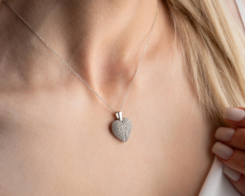 May include: A silver necklace with a heart-shaped pendant. The pendant is covered in small, sparkling stones, and the chain is delicate. The necklace is worn around the neck, and the background is blurred.