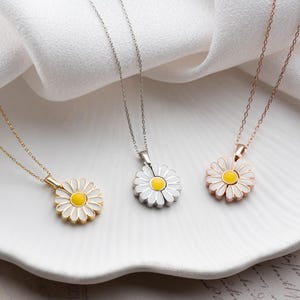 Puede incluir: Tres collares con colgante de margarita en oro, plata y oro rosa. Cada collar presenta un colgante de margarita con p&eacute;talos blancos y un centro amarillo, suspendido de una delicada cadena. Los collares se exhiben sobre una superficie blanca y ondulada.