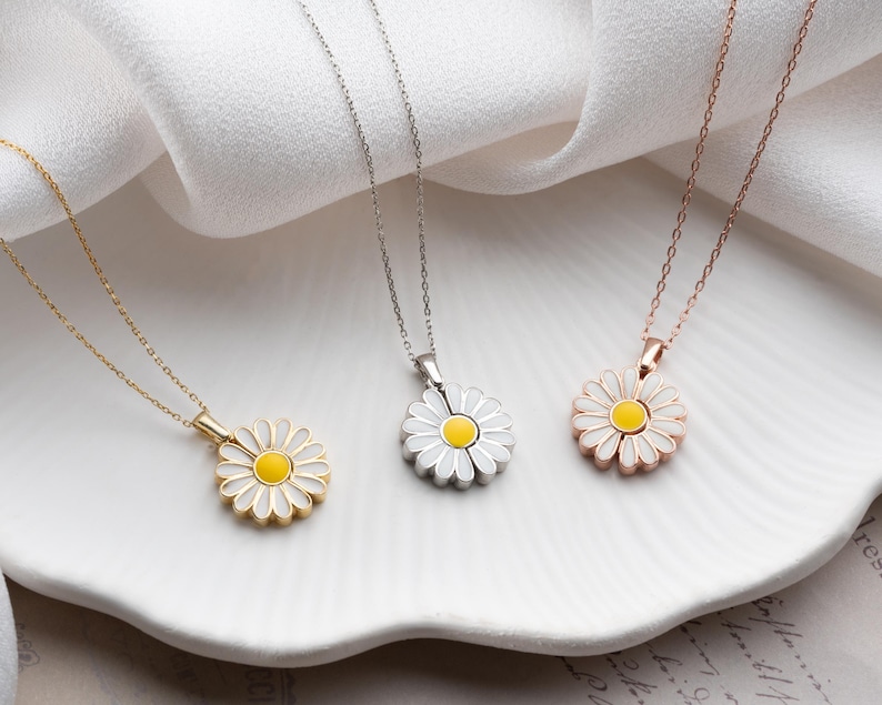 Puede incluir: Tres collares con colgante de margarita en oro, plata y oro rosa. Cada collar presenta un colgante de margarita con p&eacute;talos blancos y un centro amarillo, suspendido de una delicada cadena. Los collares se exhiben sobre una superficie blanca y ondulada.