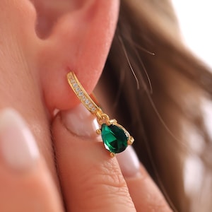 May include: A gold-colored earring with a teardrop-shaped, emerald-green gemstone. The earring is attached to a gold hoop lined with small, clear stones. The earring is worn on the ear.