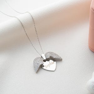 May include: A silver necklace with a heart-shaped pendant and angel wings. The heart is engraved with the name "Sophia". The wings are encrusted with small, sparkling stones. The necklace is displayed on a white, textured surface.