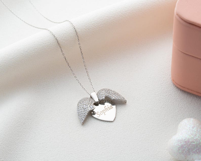 May include: A silver necklace with a heart-shaped pendant and angel wings. The heart is engraved with the name "Sophia". The wings are encrusted with small, sparkling stones. The necklace is displayed on a white, textured surface.