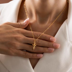 May include: A gold-colored necklace featuring a small angel pendant. The delicate chain and pendant are displayed against a white background. The angel pendant has detailed wings and a flowing garment.