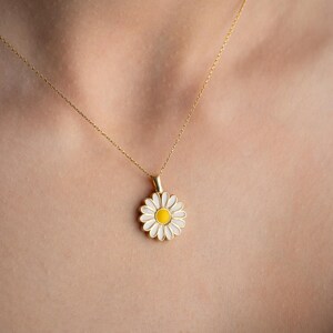 Puede incluir: Un collar dorado con un colgante de margarita. La margarita tiene p&eacute;talos blancos y un centro amarillo. El collar se lleva alrededor del cuello.