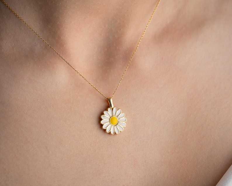 Puede incluir: Un collar dorado con un colgante de margarita. La margarita tiene p&eacute;talos blancos y un centro amarillo. El collar se lleva alrededor del cuello.