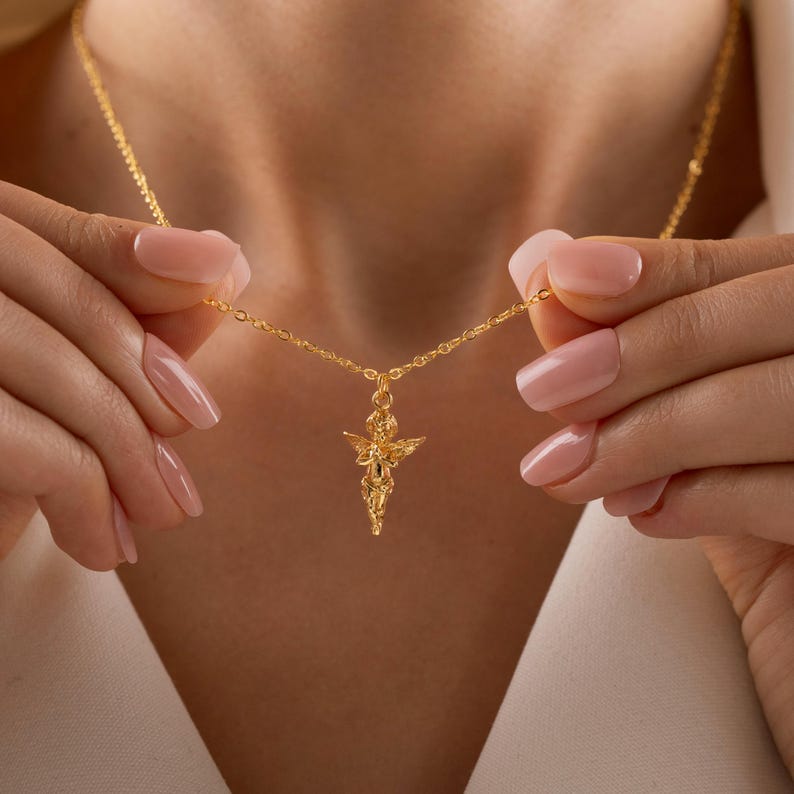 May include: A gold-colored necklace featuring a pendant in the shape of a small angel. The angel pendant has detailed wings and is suspended from a delicate chain. The necklace is being held up by a person.