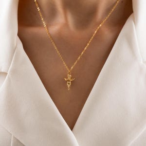 May include: A gold-colored necklace with a pendant shaped like a small angel. The angel has outstretched wings and is suspended from a delicate chain. The necklace is worn against a white shirt.