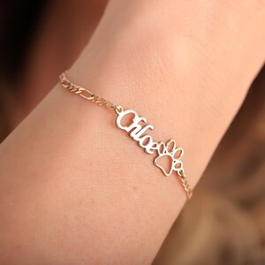 May include: A gold-coloured bracelet featuring the name "Chloe" in cursive and a paw print charm. The bracelet is worn on a wrist, highlighting the fine chain and design. This is a personalised pet-themed accessory.
