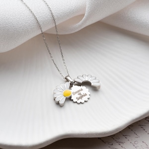 Puede incluir: Un collar de plata con tres colgantes de margaritas. Un colgante de margarita tiene un centro amarillo y p&eacute;talos blancos. El segundo colgante es una media margarita. El tercer colgante es un colgante circular con los nombres "Emma" y "Sophia" grabados.