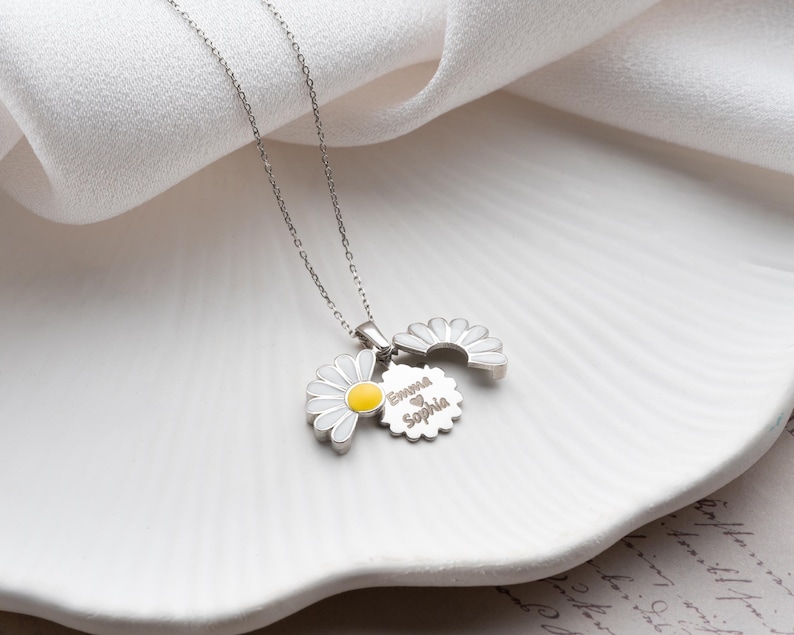 Puede incluir: Un collar de plata con tres colgantes de margaritas. Un colgante de margarita tiene un centro amarillo y p&eacute;talos blancos. El segundo colgante es una media margarita. El tercer colgante es un colgante circular con los nombres "Emma" y "Sophia" grabados.