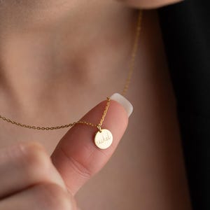 May include: A delicate gold necklace featuring a small, round pendant with the name "rachel" inscribed in cursive script. The pendant hangs from a fine chain, creating a minimalist and elegant look. The necklace is shown on a person's finger.