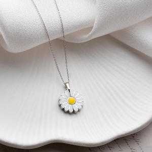 Puede incluir: Un collar con colgante de margarita plateada. El colgante presenta una margarita de esmalte blanco con un centro amarillo. El collar se muestra sobre una superficie blanca texturizada. La cadena es delicada y plateada.