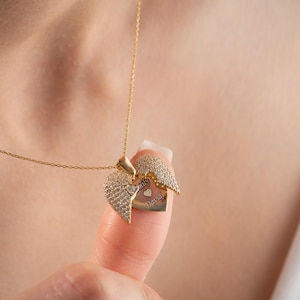 May include: A gold-colored heart-shaped pendant necklace. The pendant opens to reveal a smaller heart with engraved text. The outer heart is encrusted with small, clear stones. The necklace is displayed against a blurred background.