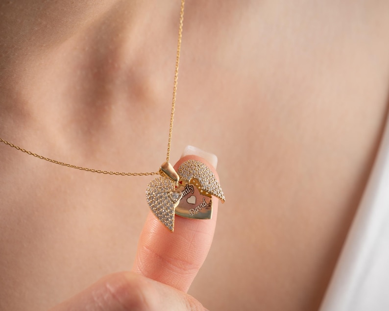 May include: A gold-colored heart-shaped pendant necklace. The pendant opens to reveal a smaller heart with engraved text. The outer heart is encrusted with small, clear stones. The necklace is displayed against a blurred background.