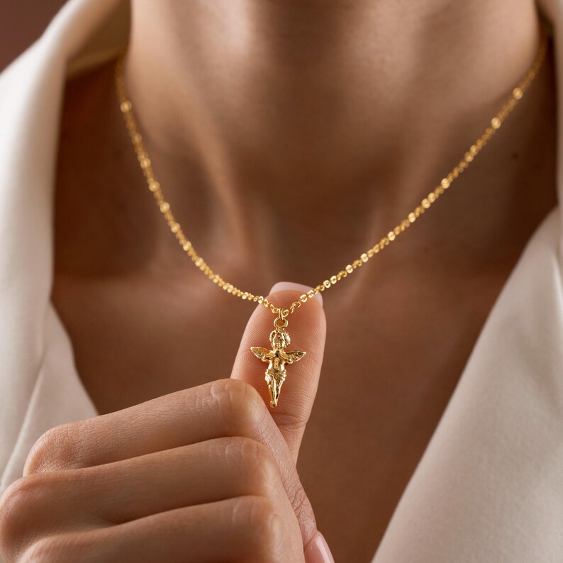 May include: A gold-colored necklace featuring a small pendant of an angel with outstretched wings. The delicate chain is visible against a white background. The pendant is held by a person's finger.