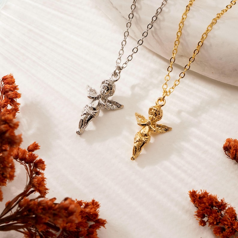 May include: Two necklaces with cherub pendants. One necklace is silver-toned, the other is gold-toned. The cherub pendants have detailed wings and are suspended from delicate chain necklaces. The background is a light, neutral color with dried orange flowers.
