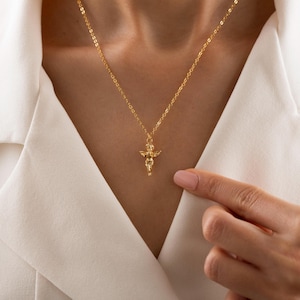 May include: A gold-colored necklace featuring a small cherub pendant. The cherub has outstretched wings and is suspended from a delicate chain. The necklace is worn against a white blouse.