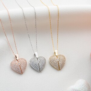 May include: Three heart-shaped pendant necklaces in rose gold, silver, and gold. Each pendant is encrusted with small, sparkling stones and features a smaller heart detail at the center. The necklaces hang on delicate chains, displayed on a white surface.