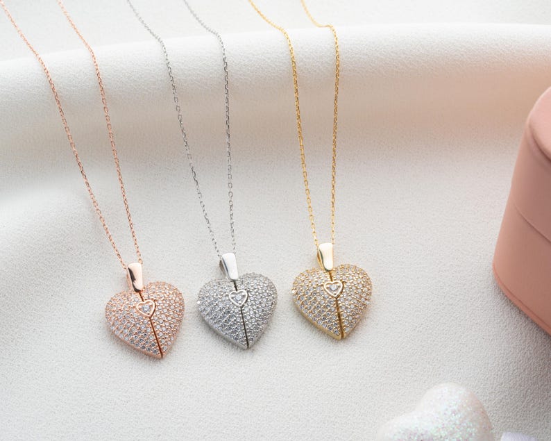 May include: Three heart-shaped pendant necklaces in rose gold, silver, and gold. Each pendant is encrusted with small, sparkling stones and features a smaller heart detail at the center. The necklaces hang on delicate chains, displayed on a white surface.