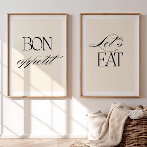 May include: Two framed art prints with the phrases "BON appétit" and "Let's EAT" in black script. The prints have a beige background and are in light brown wooden frames. A woven basket with a blanket is in front of the prints.