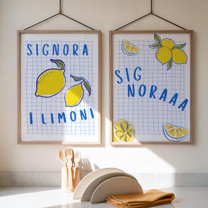 May include: Two framed art prints with lemon illustrations and Italian text. The prints feature yellow lemons on a blue grid background. Below, a set of beige plates, wooden utensils, and a folded orange cloth are arranged on a white countertop.