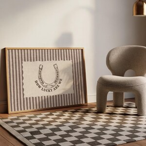 May include: A rectangular framed wall art piece featuring a vertical brown and white striped background with a central cream-colored patch. The patch displays a black line drawing of a horseshoe and the text "HOW LUCKY ARE WE" in black.
