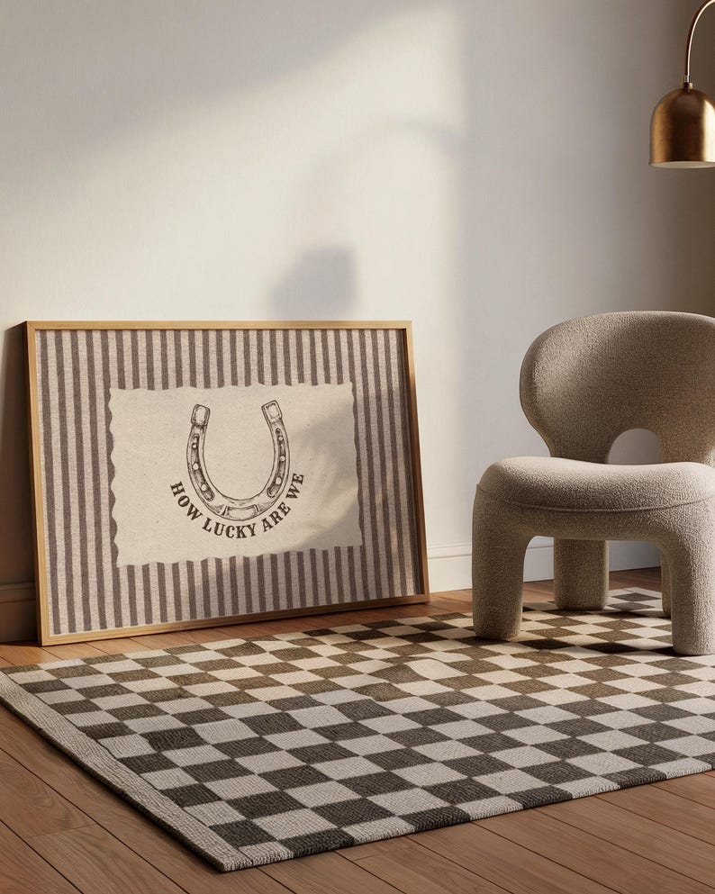 May include: A rectangular framed wall art piece featuring a vertical brown and white striped background with a central cream-colored patch. The patch displays a black line drawing of a horseshoe and the text "HOW LUCKY ARE WE" in black.