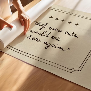 May include: A rectangular beige poster featuring a dark brown border and five stars above the text "chef was cute, would eat here again." The poster is partially rolled up, displaying a textured surface with clear, dark brown cursive lettering.