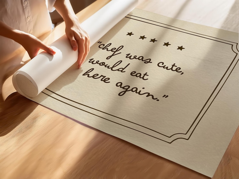 May include: A rectangular beige poster featuring a dark brown border and five stars above the text "chef was cute, would eat here again." The poster is partially rolled up, displaying a textured surface with clear, dark brown cursive lettering.