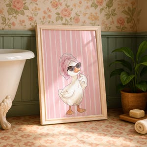 May include: A framed wall art print featuring a white duck wearing black sunglasses and a pink towel wrapped around its head. The background of the illustration consists of vertical white stripes on a soft pink field, presented in a light-colored frame.