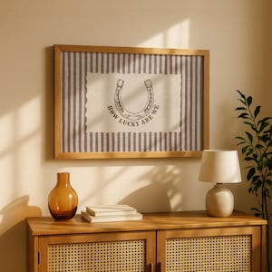 May include: A rectangular wall art piece in a light wood frame featuring a vertical grey and white striped background. Centered is a cream-colored rectangular patch with a scalloped edge, displaying a horseshoe illustration and the text HOW LUCKY ARE WE.