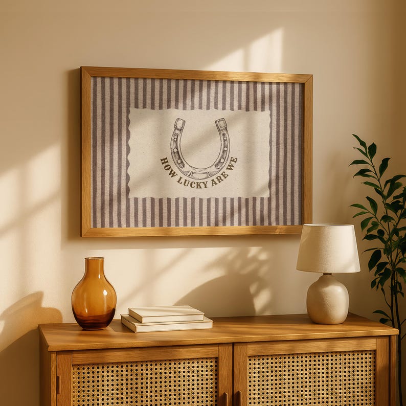 May include: A rectangular wall art piece in a light wood frame featuring a vertical grey and white striped background. Centered is a cream-colored rectangular patch with a scalloped edge, displaying a horseshoe illustration and the text HOW LUCKY ARE WE.