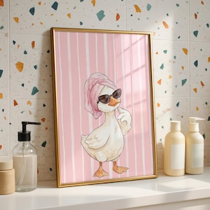 May include: A framed wall art print featuring a cartoon duck wearing a pink towel wrapped around its head and black sunglasses. The duck is positioned against a background of vertical pink and white stripes, rendered in a watercolor style.