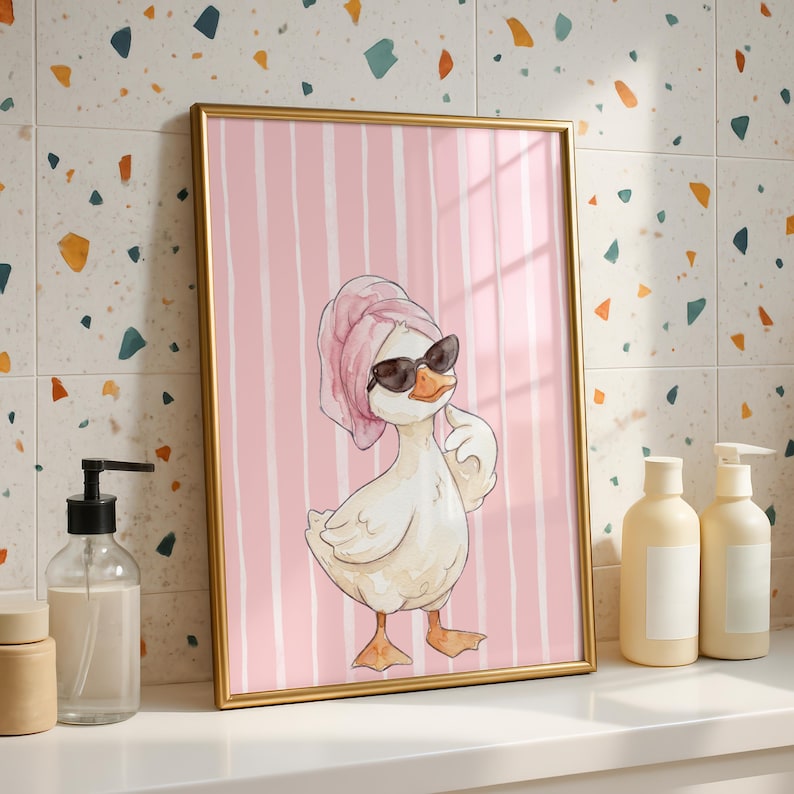 May include: A framed wall art print featuring a cartoon duck wearing a pink towel wrapped around its head and black sunglasses. The duck is positioned against a background of vertical pink and white stripes, rendered in a watercolor style.
