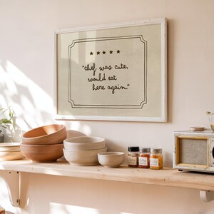 May include: A rectangular wall art print in a white frame featuring a beige background with a black border, five stars, and the text "chef was cute, would eat here again." written in a handwritten script font centered within the frame.