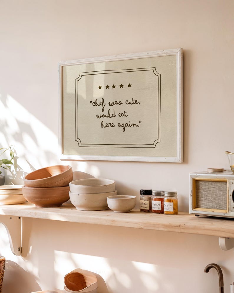 May include: A rectangular wall art print in a white frame featuring a beige background with a black border, five stars, and the text "chef was cute, would eat here again." written in a handwritten script font centered within the frame.