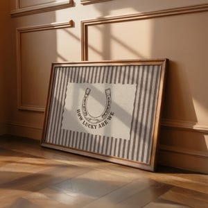 May include: A rectangular framed wall art piece featuring a beige background with vertical dark brown stripes. Centered is a cream-colored paper cutout with a horseshoe illustration and the black text "HOW LUCKY ARE WE" printed below it.