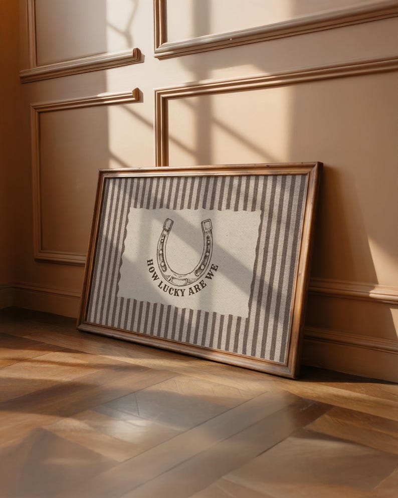 May include: A rectangular framed wall art piece featuring a beige background with vertical dark brown stripes. Centered is a cream-colored paper cutout with a horseshoe illustration and the black text "HOW LUCKY ARE WE" printed below it.