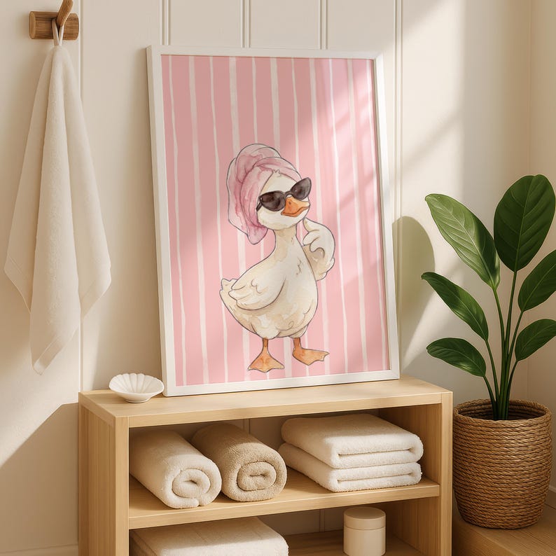 May include: A framed wall art print featuring a white duck wearing black sunglasses and a pink towel wrapped around its head. The background of the illustration consists of vertical pink and white stripes. The duck is standing in a pose.