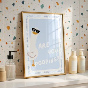 May include: A framed print featuring a cartoon goose wearing sunglasses, set against a light blue background. The text "ARE YOU POOPING?" is displayed. The frame is gold, and the print is displayed on a white surface.