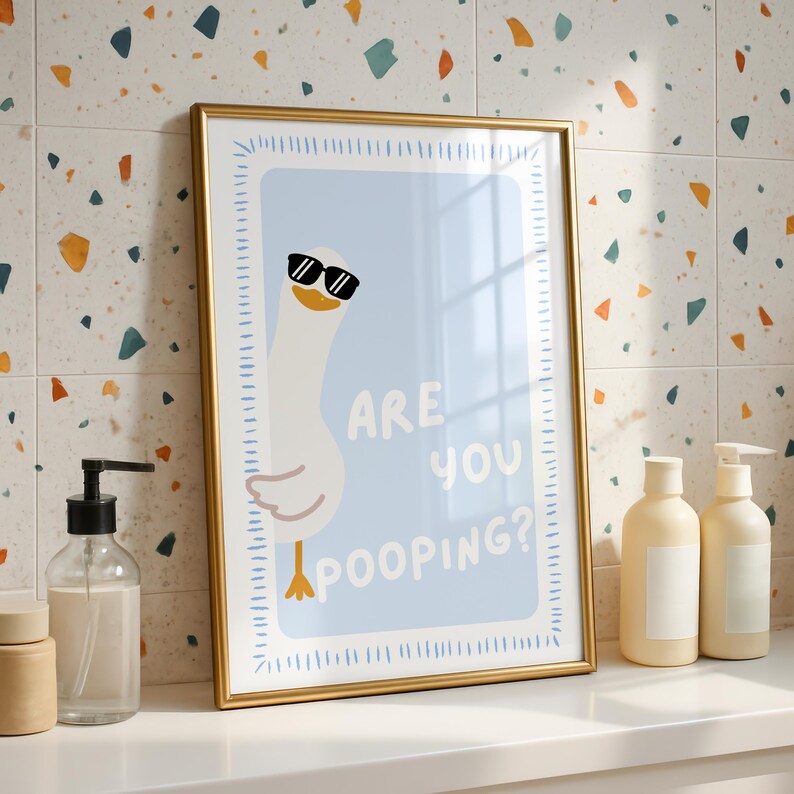 May include: A framed print featuring a cartoon goose wearing sunglasses, set against a light blue background. The text "ARE YOU POOPING?" is displayed. The frame is gold, and the print is displayed on a white surface.