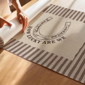 May include: A rectangular wall art print featuring a central cream-colored panel with a black line-art illustration of a horseshoe and the text "HOW LUCKY ARE WE" printed below it, framed by a brown and white striped border on all sides.