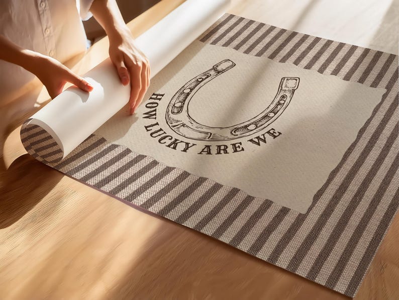 May include: A rectangular wall art print featuring a central cream-colored panel with a black line-art illustration of a horseshoe and the text "HOW LUCKY ARE WE" printed below it, framed by a brown and white striped border on all sides.