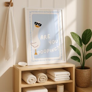 May include: A framed print with a cartoon goose wearing sunglasses and the text "ARE YOU POOPING?" is displayed above a wooden shelf with rolled and folded towels. A white towel hangs on a wooden hook, and a potted plant sits to the right.