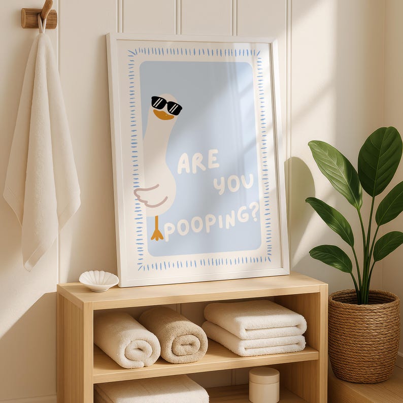 May include: A framed print with a cartoon goose wearing sunglasses and the text "ARE YOU POOPING?" is displayed above a wooden shelf with rolled and folded towels. A white towel hangs on a wooden hook, and a potted plant sits to the right.