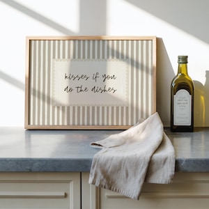 May include: A framed print with the text "kisses if you do the dishes" sits on a countertop next to a bottle of olive oil and a folded linen towel. The print has a striped background.