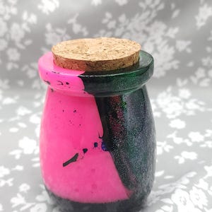 Puede incluir: Un pequeño tarro decorativo con tapón de corcho. El tarro está pintado con un vibrante diseño dividido en rosa intenso y verde oscuro, con una base negra. El tapón de corcho es de color marrón claro.