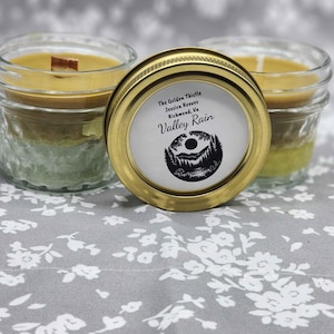 Pode incluir: Três velas em frascos de vidro com um design de cera em camadas e pavios de madeira. As velas estão em frascos transparentes com uma tampa dourada que tem o texto "Valley Rain" e uma cena de montanha. As velas estão sobre uma superfície cinza com estampa floral.