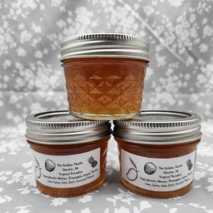 May include: Three small glass jars of tropical fruit jam with silver lids are stacked. The top jar is on top of two jars. Each jar has a label with text and illustrations of a coconut, mango, and pineapple. The jam is a golden-amber color.