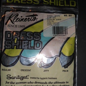 Puede incluir: Un paquete vintage de protectores de axilas Kleinert's Dress Shields. El empaque muestra ilustraciones de diferentes formas de protectores: Regular, Crescent, Jiffy y Pin-In. El texto incluye "Sanitized" y "100% Algodón forrado de vinilo."